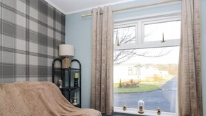 A room with a bed covered in a beige blanket, a window with curtains, a black shelf with decorative items, and a view of a house and lawn outside. - Beechtree Lodge 55 (Denny)