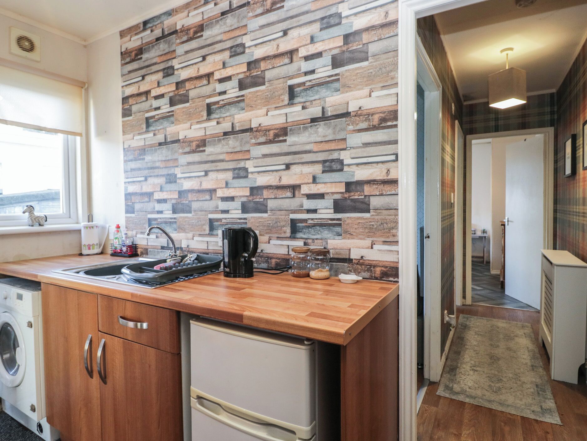 Cocina compacta con encimera de madera, lavaplatos integrado y refrigerador. Una pared decorativa con diferentes texturas de madera se encuentra detrás de la encimera.