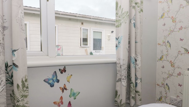 A bedroom with a window, white curtains with a floral pattern, and butterfly decals on the wall. - Beechtree Lodge 46 (Denny)
