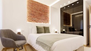 A modern bedroom with a brick accent wall, a large bed with white bedding, a grey armchair, and a bedside table with a lamp. - Genteel Home Concha Espina I (Madrid)