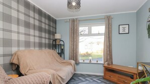 A bedroom with a bed covered in a beige blanket, a wooden nightstand, a lamp, and a window with curtains. - Beechtree Lodge 56 (Denny)