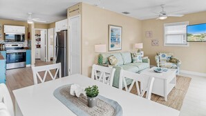 A modern kitchen with white cabinets, a stainless steel refrigerator, and a built-in oven. There is a dining area with a white table and chairs, and a living room with a light blue sofa and a patterned armchair. - Lou's Starfish Haven - Beach Front (Indian Rocks Beach)