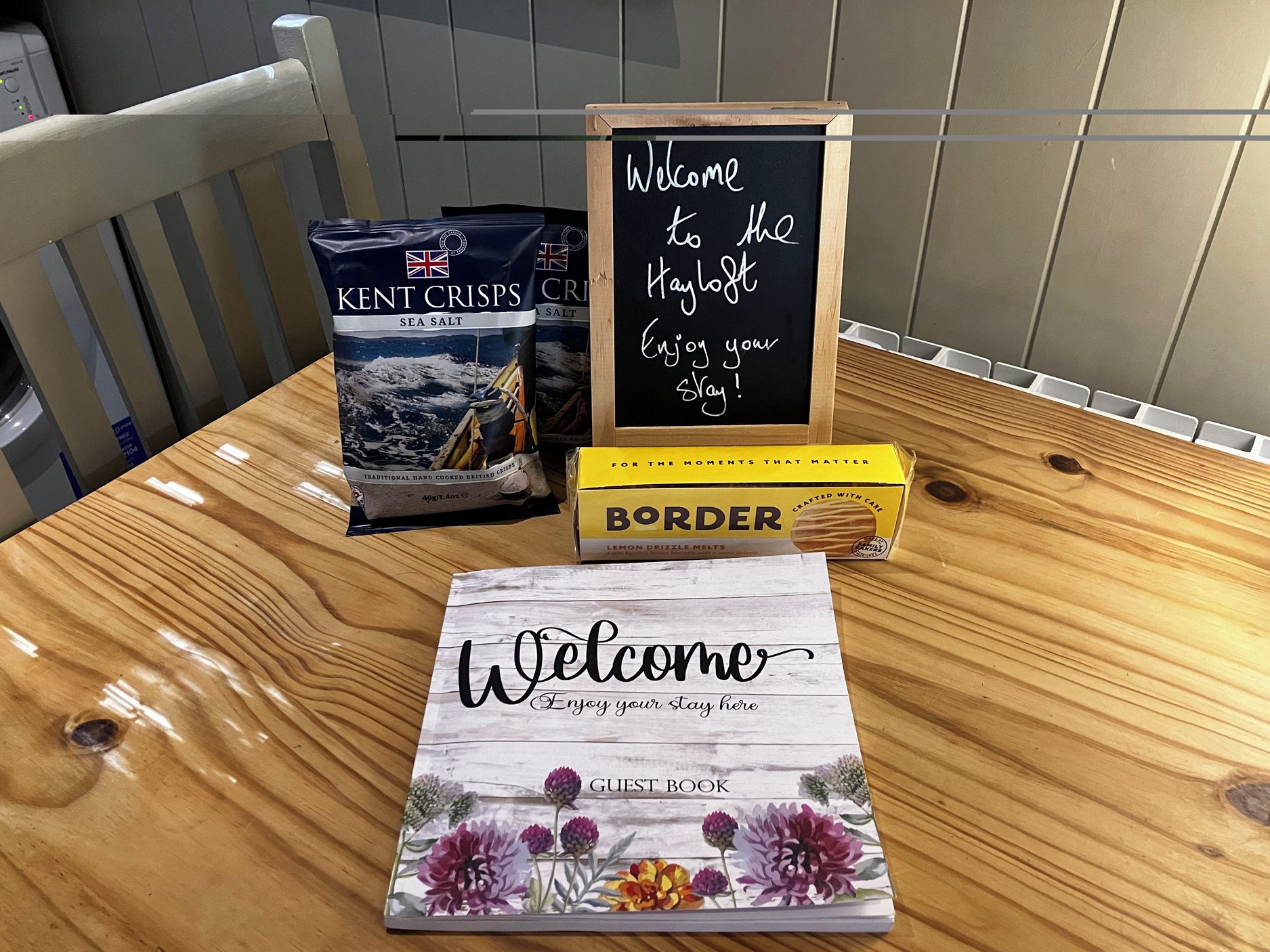 A wooden table with a welcome sign, a guest book, and a box of lemon drizzle melts.