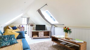 A cozy loft-style living room with a sloped ceiling, a large window, a blue sofa, a wooden coffee table, and a television. - The Hayloft - Beautiful Barn Stay (Ashford)