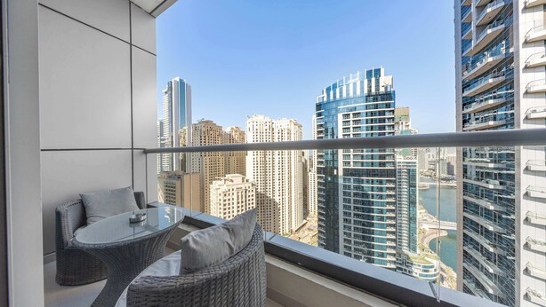A balcony with a glass table and wicker chairs, overlooking a cityscape with high-rise buildings and a body of water. - StayShort - 2BR in Dubai Marina that Sleeps 6 with Water Views! (Dubai Marina)