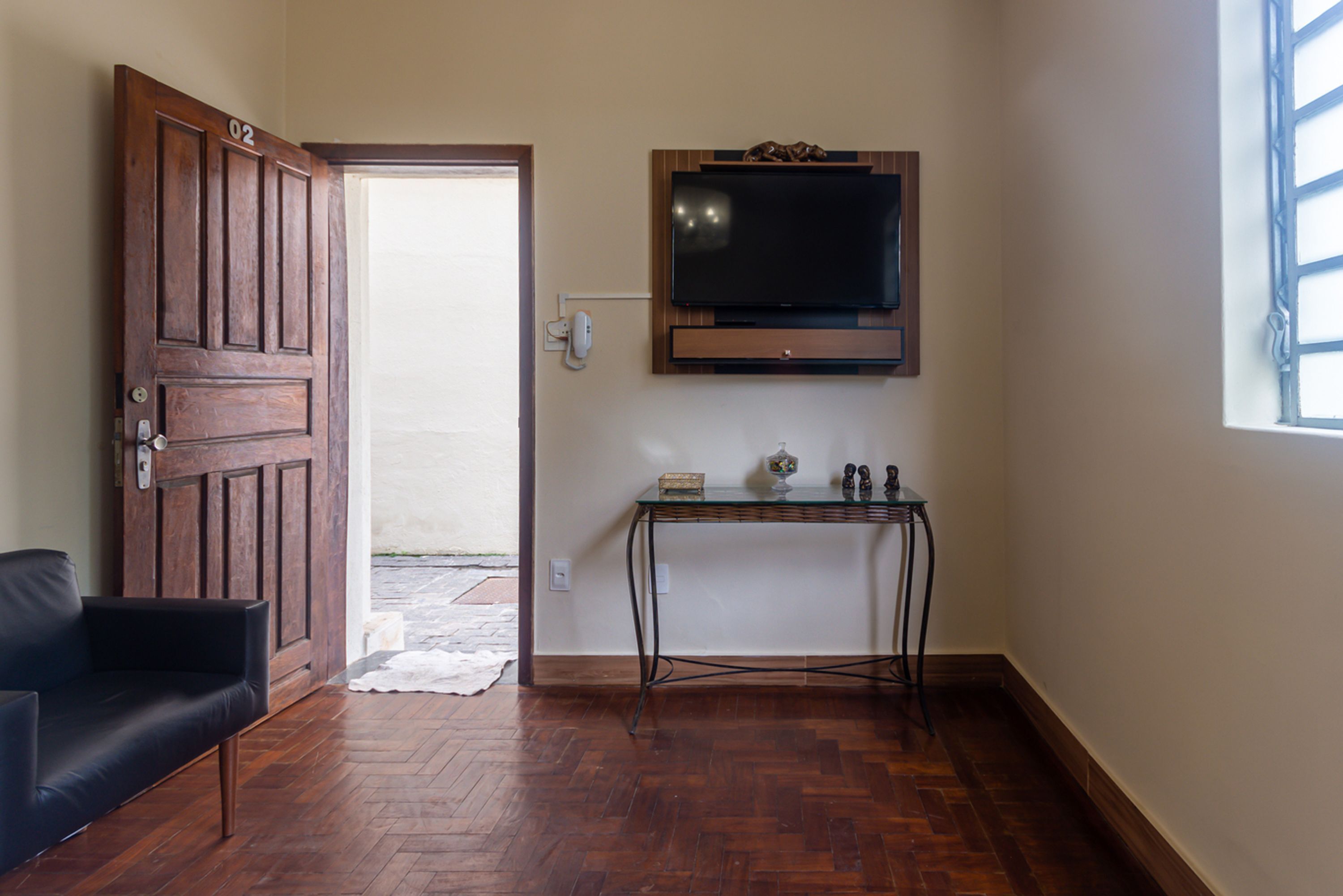 A room with a wooden door, a black sofa, a television mounted on the wall, and a small table with decorative items.