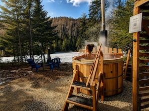 A hot tub in a forested area with a wooden deck and chairs. - Aurora | Lakefront | Hot tub | Fireplace (Matawinie Regional County Municipality)