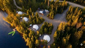 An aerial view of a lakeside cabin complex with yurts, a main lodge, and a dock with kayaks.