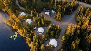 Luftaufnahme eines Chaletkomplexes am Seeufer mit Jurten, einem Hauptgebäude und einem Steg mit Kajaks.