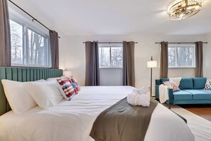 A hotel room with a large bed, a sofa, and a lamp. There are windows with curtains, a ceiling light, and a view of trees outside. - Adventure Peak Lodge • Luxe Game Room Getaway (Tobyhanna)