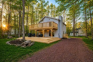 A two-story cabin with a deck, surrounded by trees and a gravel driveway. - Adventure Peak Lodge • Luxe Game Room Getaway (Tobyhanna)