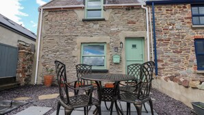 A stone building with a green door and windows, a round table with chairs, and a gravel ground.