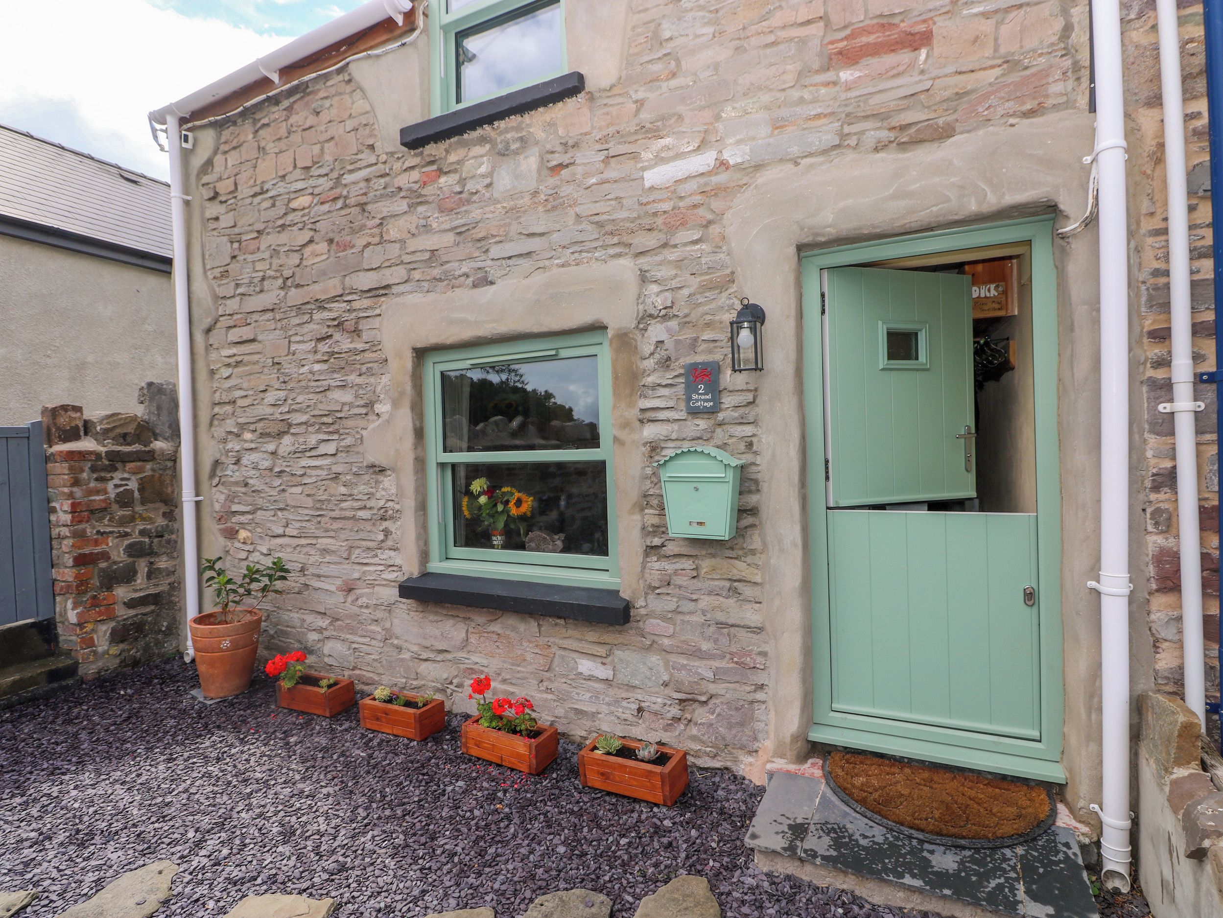 Un bâtiment en pierre avec une porte verte, une fenêtre ornée de fleurs et une enseigne indiquant « 2 Strand Cottage ».
