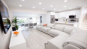 A modern living room with a sofa, a coffee table, and a TV. There is a kitchen area with appliances and cabinets.