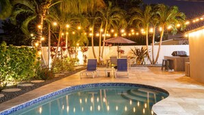 An outdoor pool area with a curved edge, surrounded by palm trees and a white fence. There are lounge chairs and a small table with an umbrella.