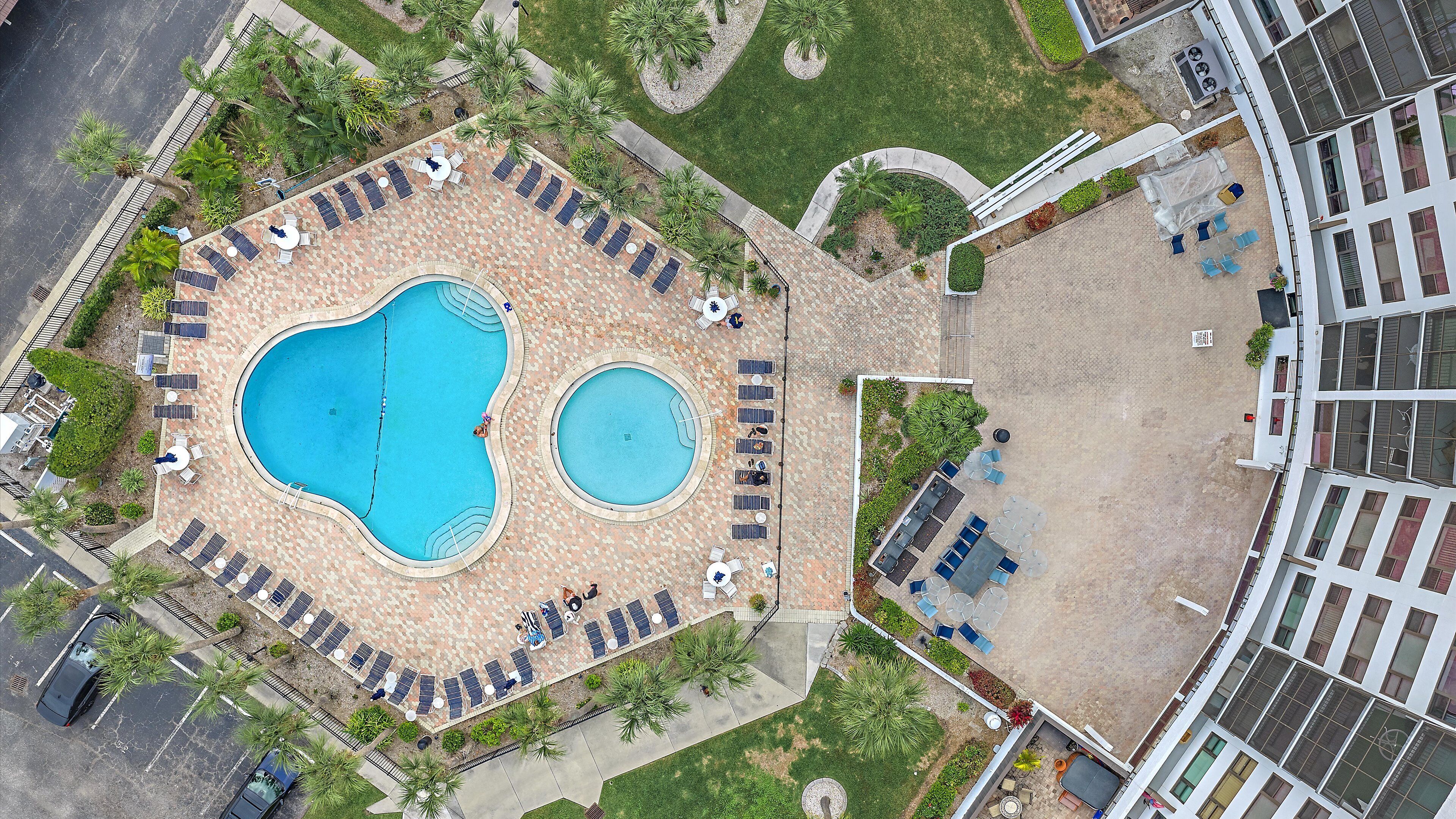 An aerial view of a resort with a large central pool, smaller adjacent pool, and multiple sun loungers and umbrellas around the pools.