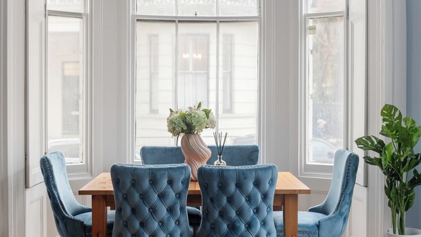 A dining area with a wooden table and four blue upholstered chairs. A vase with flowers and a candle holder are on the table.
