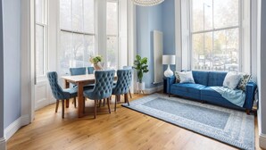 A dining area with a wooden table and blue upholstered chairs, a blue sofa, and a large window with a view of trees.