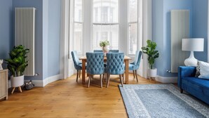 A dining area with a wooden table and blue upholstered chairs, flanked by white radiators and potted plants.
