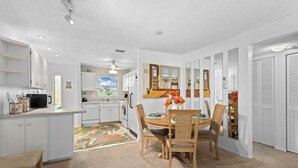Una cocina compacta con muebles blancos, un comedor con mesa y sillas de madera, y vista al salón a través de un puerta abierta.