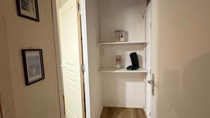A narrow hallway with a wooden floor, white walls, and a door leading to another room. There are two framed pictures on the wall and a shelf with a coffee maker and a mug.