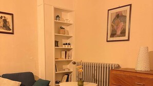 A cozy room with a sofa, a small table, a dresser, a radiator, and shelves with books and decorative items.