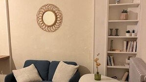 A living room with a blue sofa, a round mirror, a white bookshelf, and a small table with a vase.
