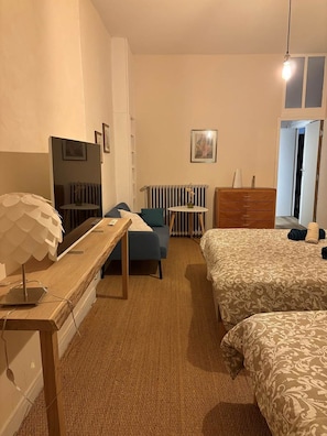 A hotel room with a bed, a desk with a lamp, a chair, a radiator, and a dresser. - THE FAMILY ROOM (Aubusson)