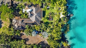 Vue aérienne d’une propriété côtière, avec une maison entourée d’une végétation luxuriante et de palmiers, située à proximité d’une mer d’un bleu limpide.