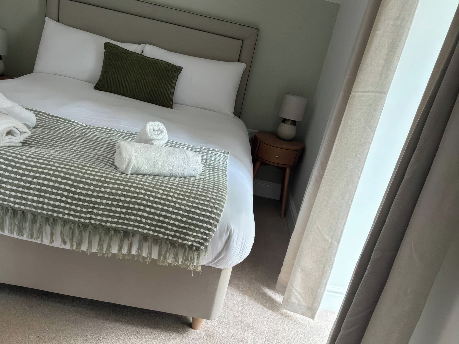 Ein ordentlich gemachtes Bett mit einer gestreiften Decke und aufgerollten Handtüchern, einem grünen Kopfkissen und einem Nachttisch mit einer Lampe.