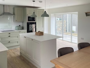 Una cocina moderna con armarios blancos, una isla central, iluminación colgante y un amplio ventanal con vistas al exterior.