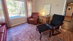 Une pièce avec une grande fenêtre, un fauteuil en cuir marron, un fauteuil noir rembourré, un meuble en bois et un tapis à motifs.