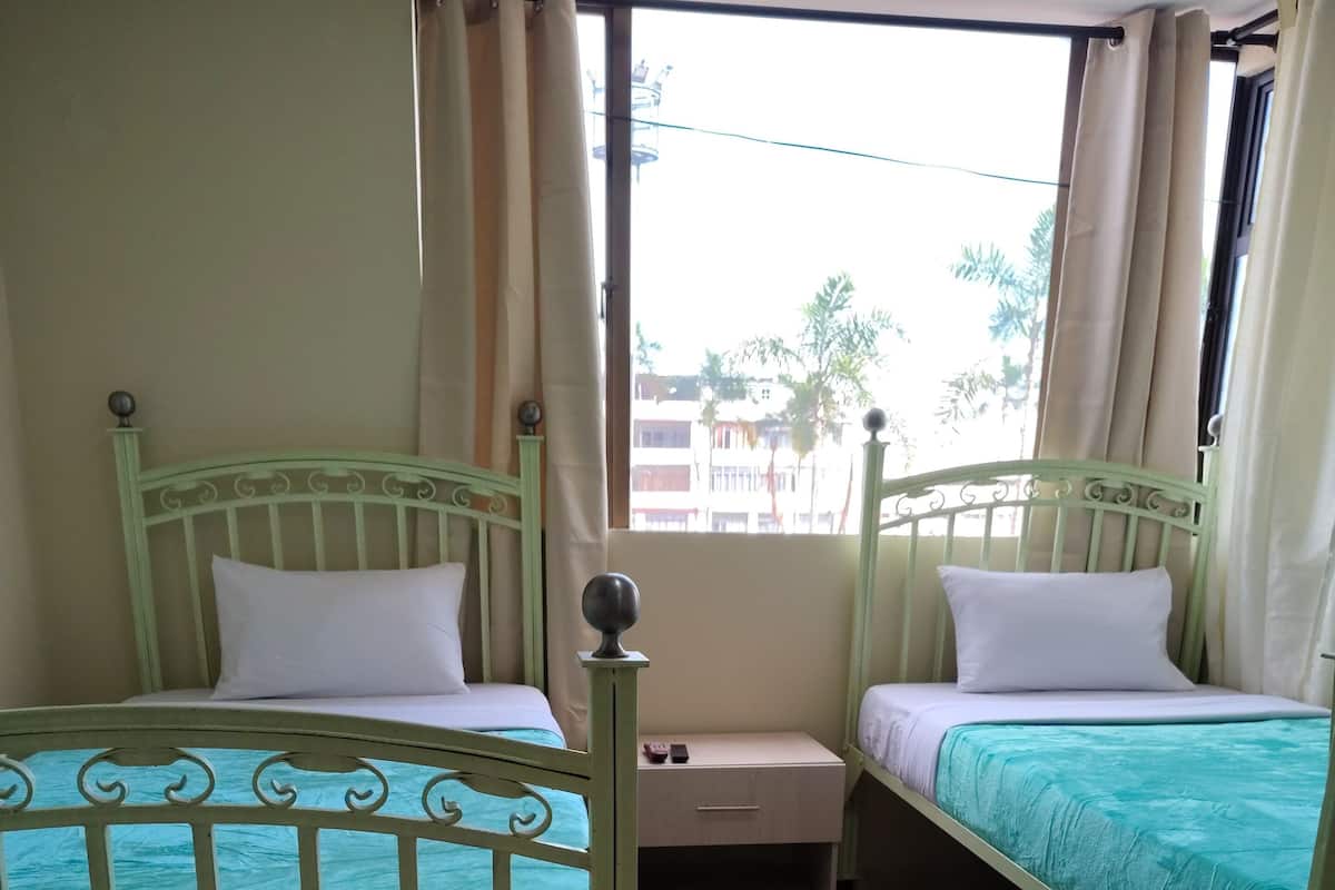 Una habitación con dos camas individuales, una mesita de noche pequeña, una ventana con cortinas y vistas a edificios.