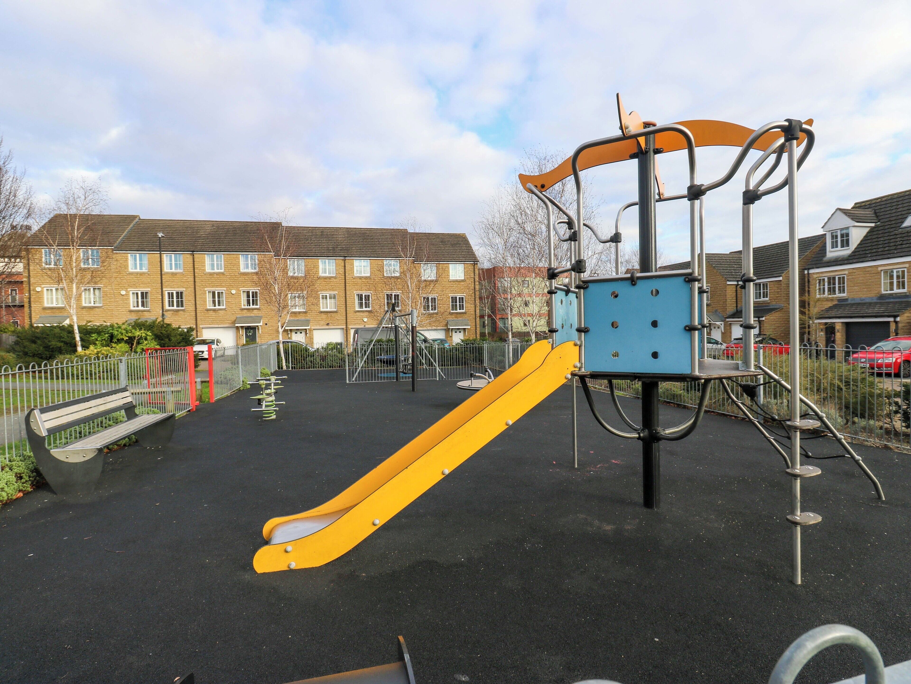 Ein Spielplatz mit einer gelben Rutsche und Klettergerüsten, umzäunt, mit Wohnhäusern im Hintergrund.
