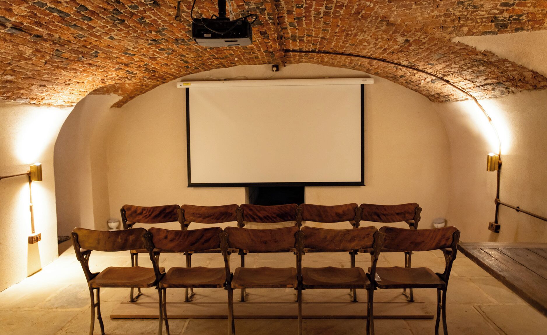 An arched room with a projector screen, wooden benches, and wall-mounted lights.