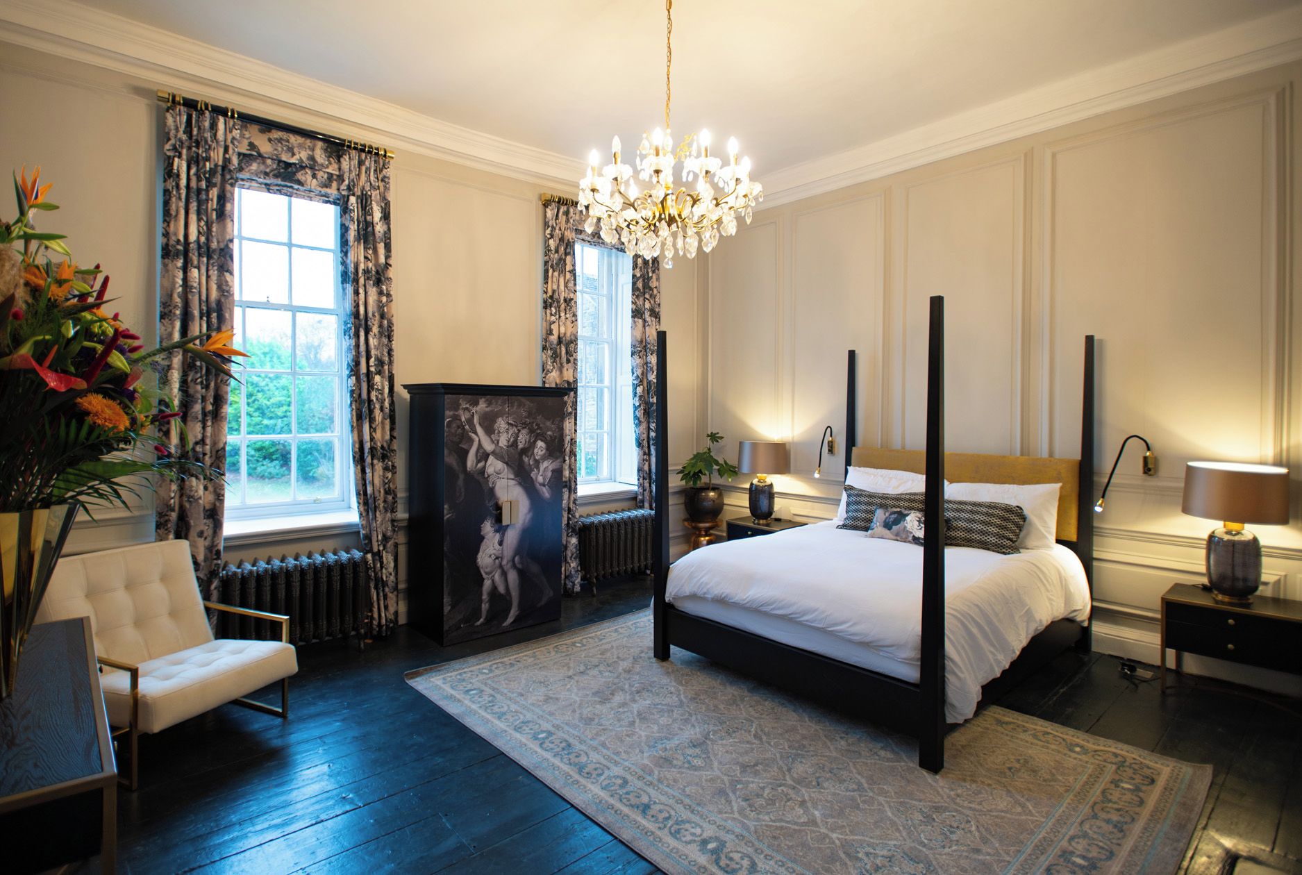 A spacious bedroom with a large bed, a sitting area with a chair, a decorative cabinet, and a view of greenery outside.