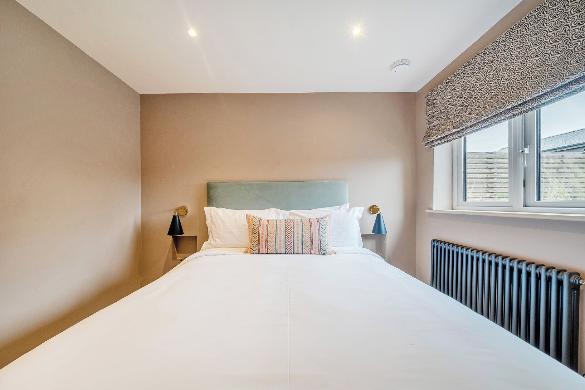 A bedroom with a bed, two bedside tables, a window with patterned curtains, and a radiator.