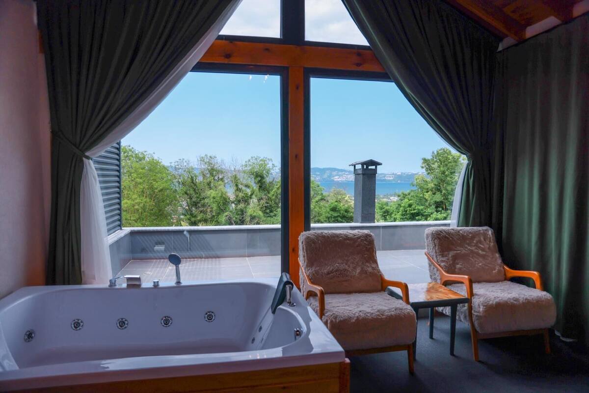 A balcony with a hot tub, two cushioned chairs, and a view of greenery and a distant structure.