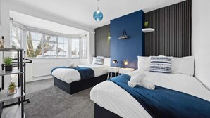 A modern bedroom with two beds, a window with blinds, a desk, and a shelf with plants.