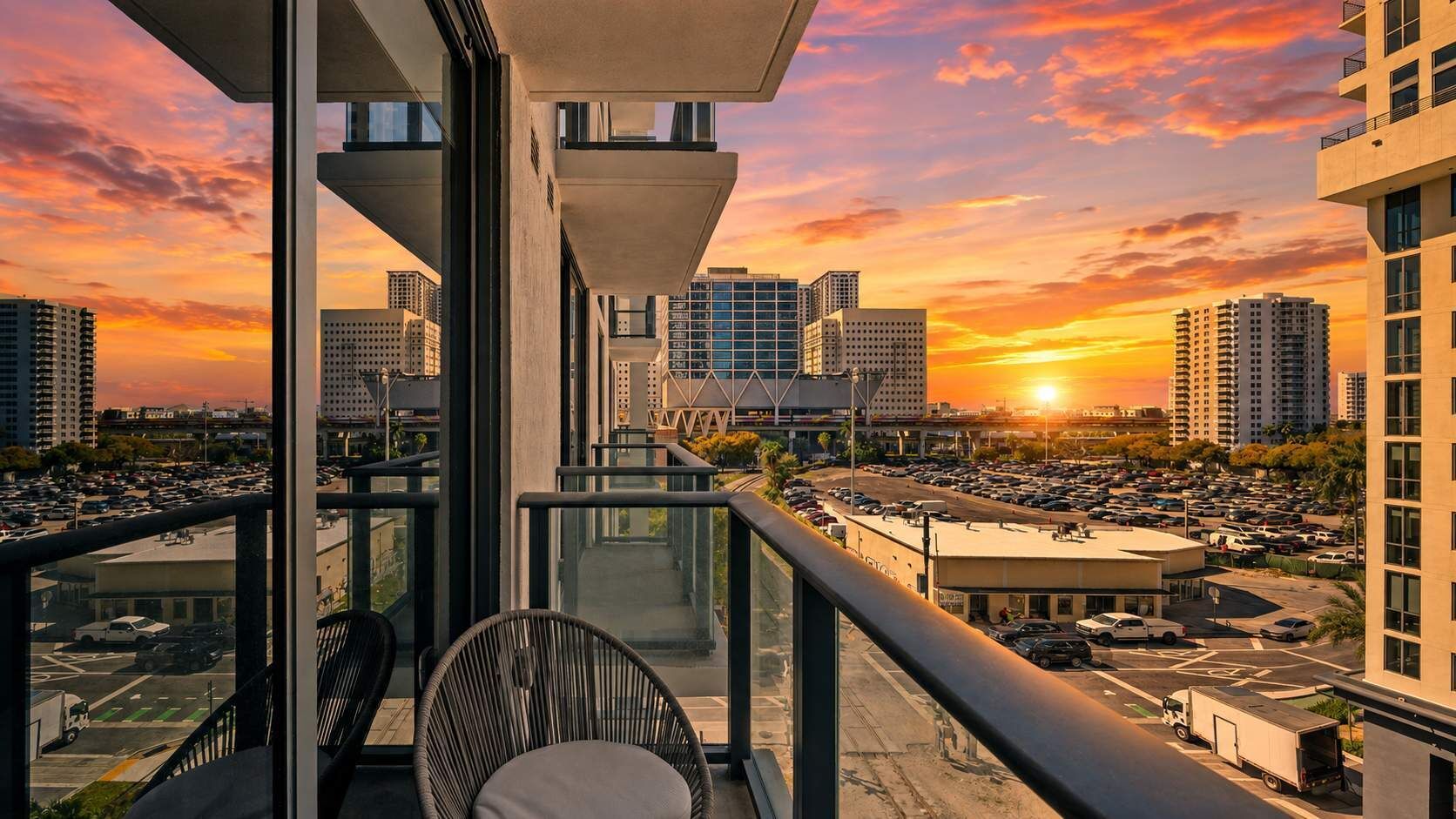 Ein Balkon mit Blick auf eine Stadtlandschaft bei Sonnenuntergang, mit modernen Gebäuden und einem Parkplatz.