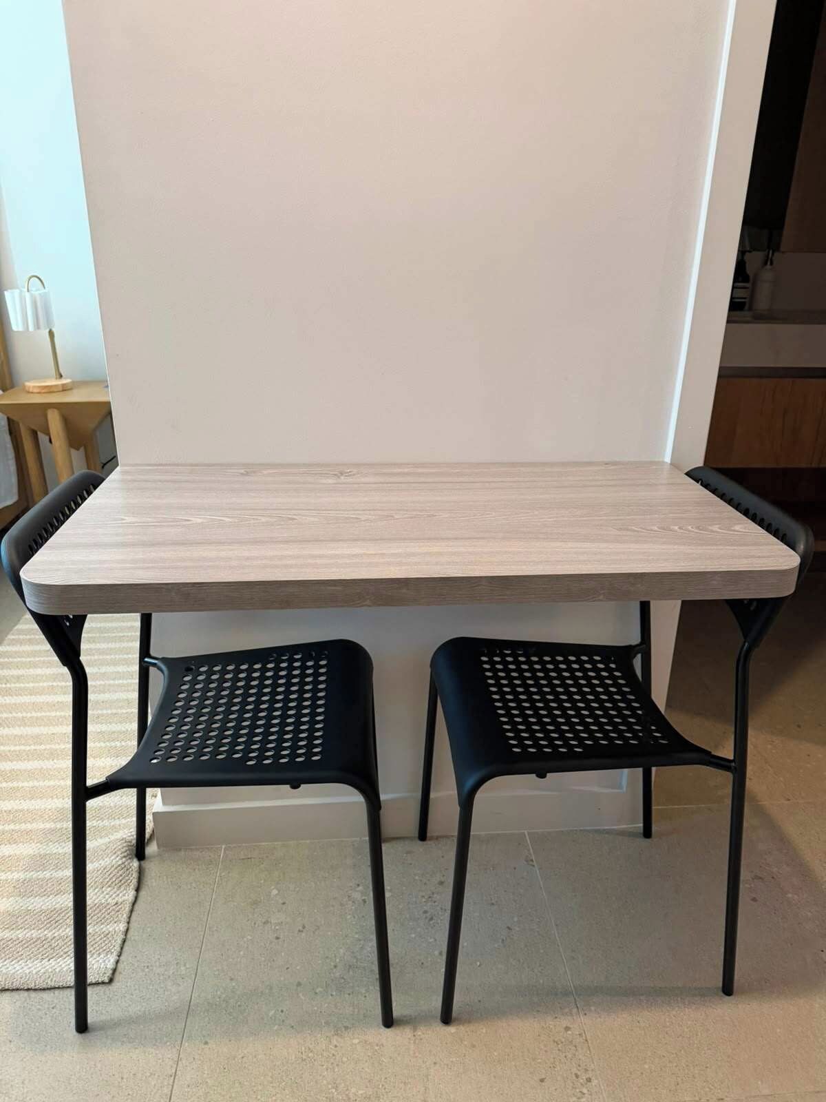 A rectangular table with a light wood finish and two black metal chairs with perforated seats.