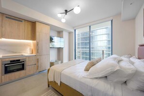 A modern bedroom with a large bed, a built-in TV, and a kitchen area with wooden cabinets and an oven. - Sleek Studio | 24/7 Concierge | Downtown (Miami)