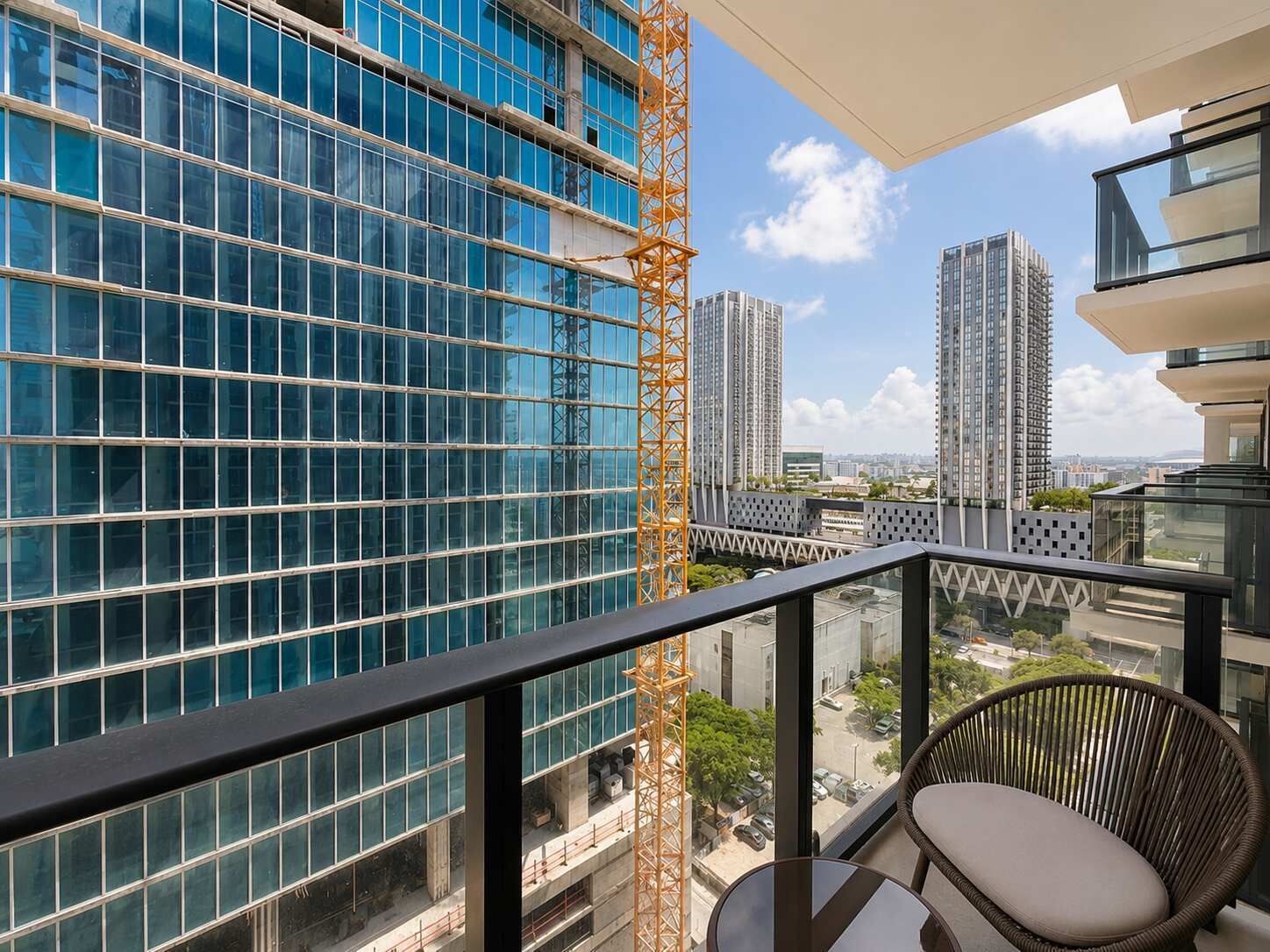 A balcony with a view of high-rise buildings, a construction crane, and a wicker chair.