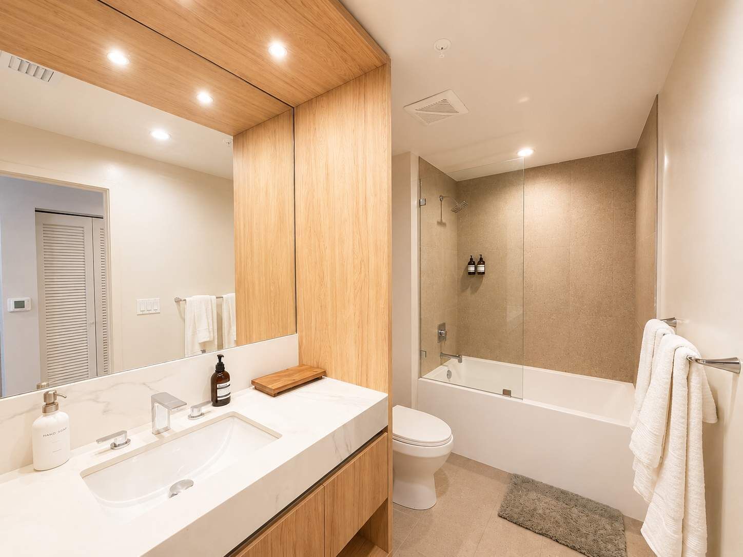 A modern bathroom with a wooden vanity, a large mirror, a bathtub, and a toilet.