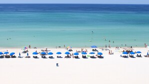 Une plage de sable blanc bordée d’une eau d’un bleu cristallin. On y trouve plusieurs parasols bleus et des chaises longues.