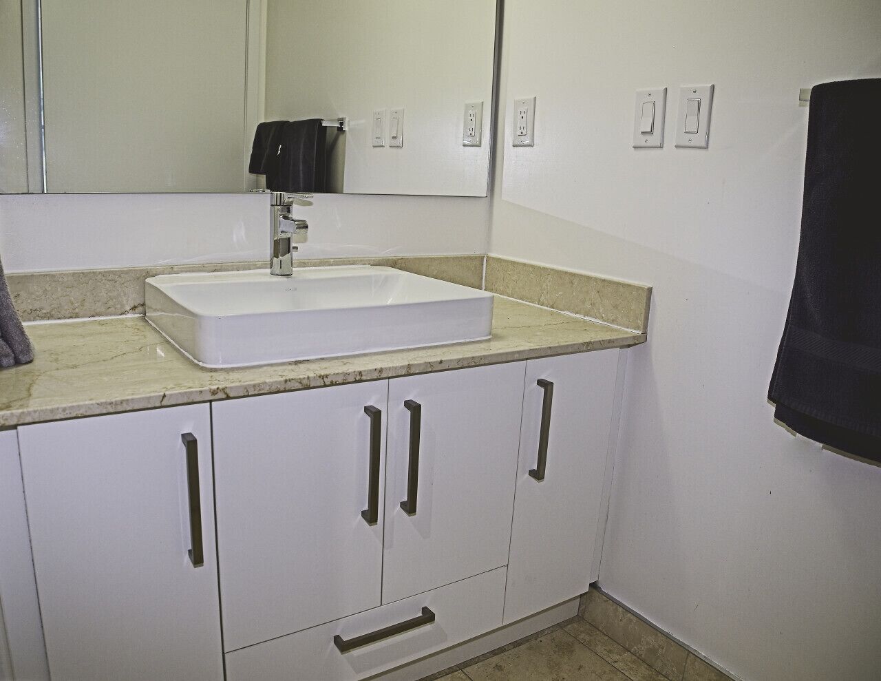 A bathroom with a white sink, beige countertop, and white cabinets with black handles. There are two light switches on the wall and a towel hanging on the right.