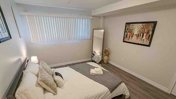 A bedroom with a bed, a window with blinds, a mirror, and a painting on the wall.