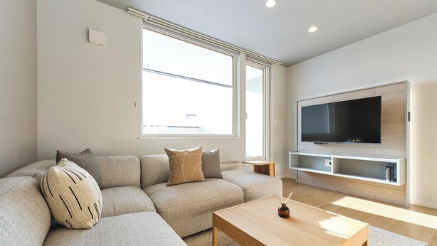 A modern living room with a sectional sofa, a wooden coffee table, a flat-screen TV mounted on a wooden wall, and a large window providing natural light. - Higashikawa Hinna (Higashikawa)