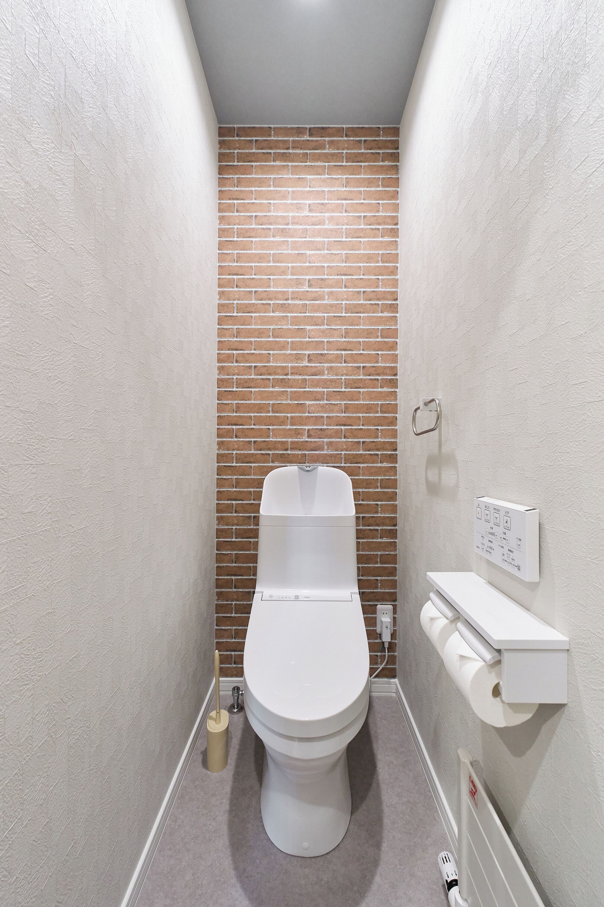 Un baño pequeño y estrecho con inodoro blanco, pared de ladrillo y estante blanco con portaescobillas.