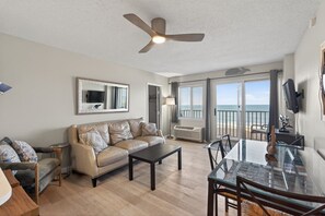 Ein Wohnzimmer mit Sofa, Couchtisch und Esstisch. Es gibt einen Deckenventilator, einen Fernseher und einen Blick auf das Meer durch große Fenster.